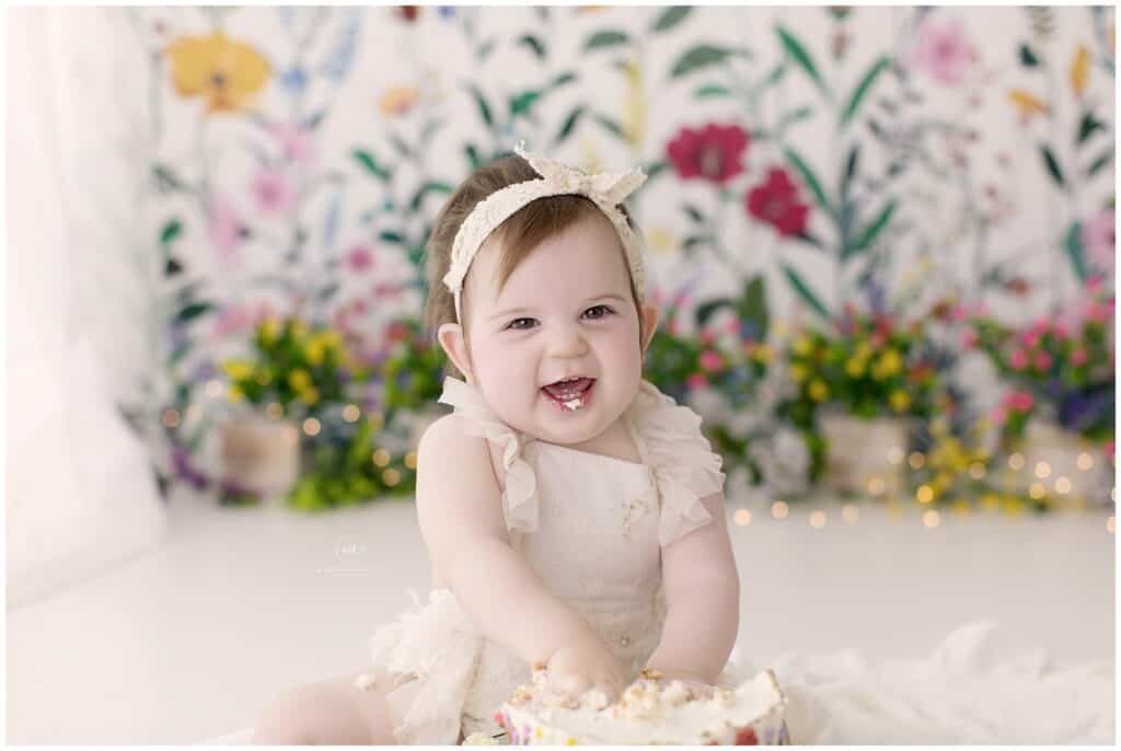 Bright smiling baby girl with a headband, celebrating her first birthday with a cake smash, in a floral background, captured by Mary Christine Photography.