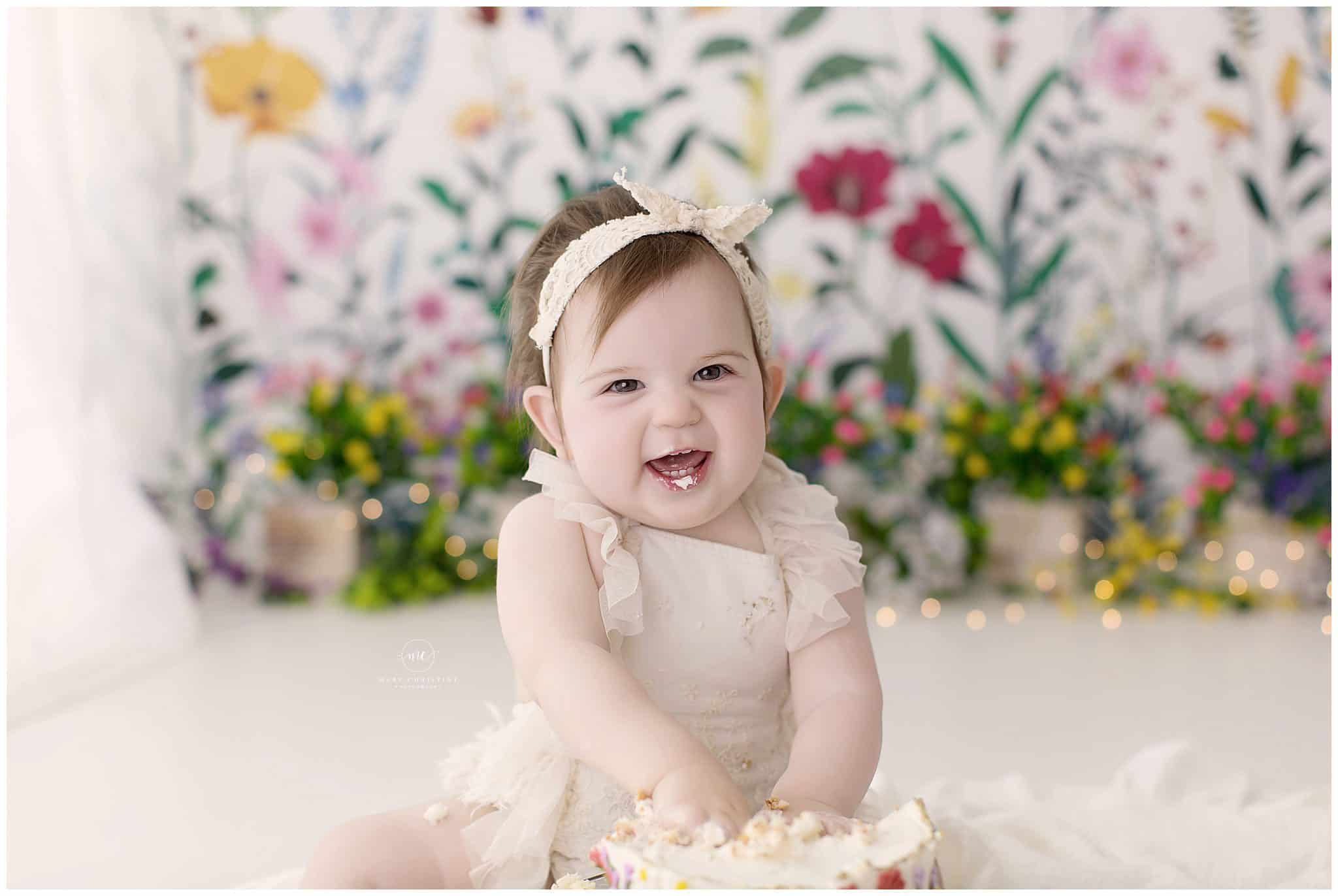 Bright smiling baby girl with a headband, celebrating her first birthday with a cake smash, in a floral background, captured by Mary Christine Photography.