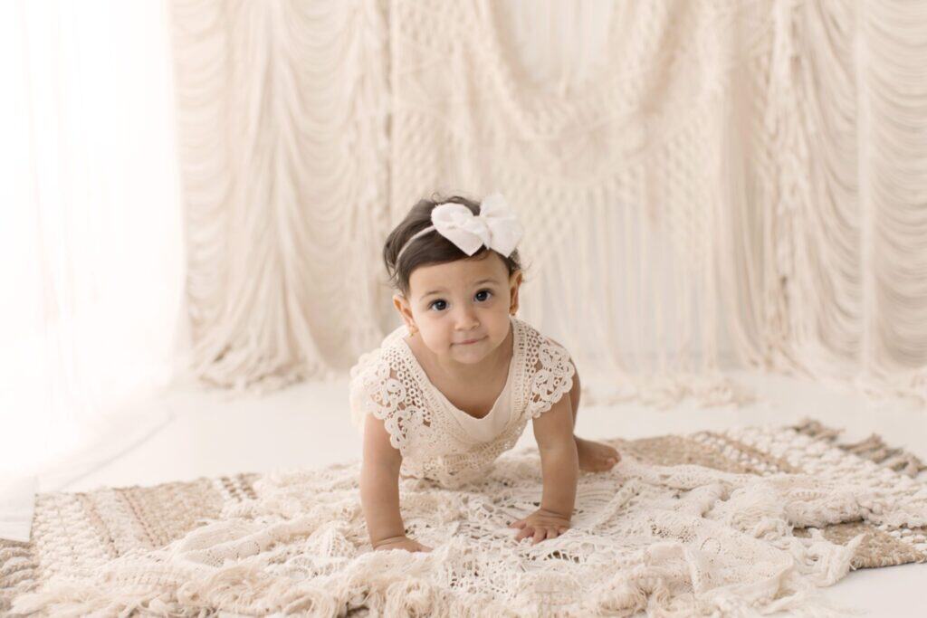 Soft-focused image of a baby girl in lace dress with a large white bow, crawling on a cream textured blanket, with elegant beige draped backdrop for timeless newborn photography.