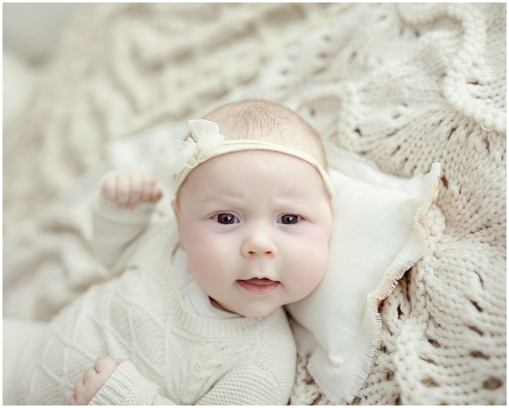 Soft-focus portrait of a baby girl in cream sweater, lying on a cozy textured knit blanket, with delicate headband, capturing adorable innocence and newborn photography artistry.