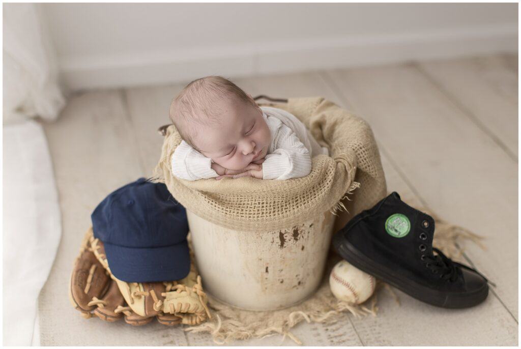 Soft focus newborn baby sleeping in a rustic bucket with baseball glove, baseball, sneakers, and a navy cap, highlighting newborn photography and family portraiture by Mary Christine Photography.