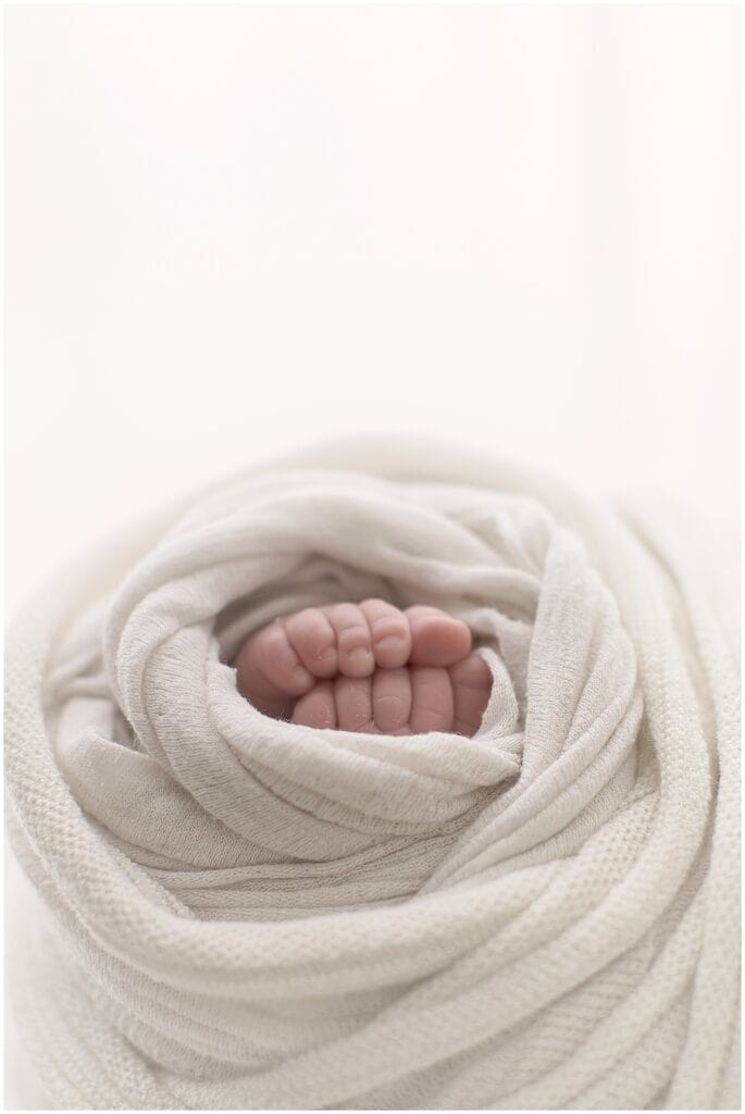 Soft newborn baby feet wrapped in cozy cream-colored swaddle blanket, focusing on delicate tiny toes, natural light, and gentle tones perfect for birth and baby photography.