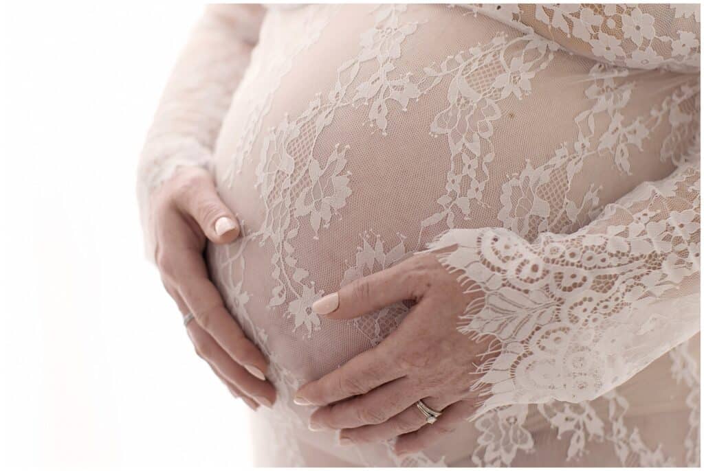 Delicate lace maternity dress highlighting intricate floral embroidery on the sheer fabric, showcasing an expecting mother’s beautiful belly with hands gently cradling it.