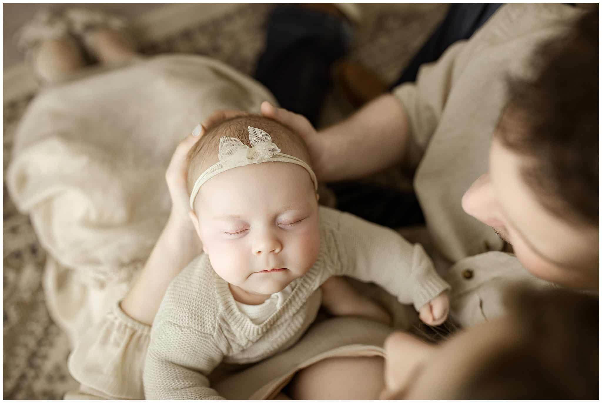 Soft-focus portrait of a newborn girl with a beige headband, peacefully sleeping on her mother’s lap, capturing intimate mother-baby bonding moments in warm, natural light, professional newborn photography.