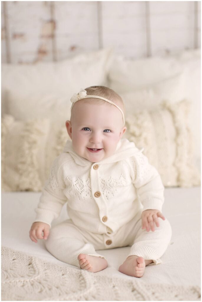 Portrait of a 6 month old baby girl sitting on a bed wearing a cream knit sweater romper and smiling.