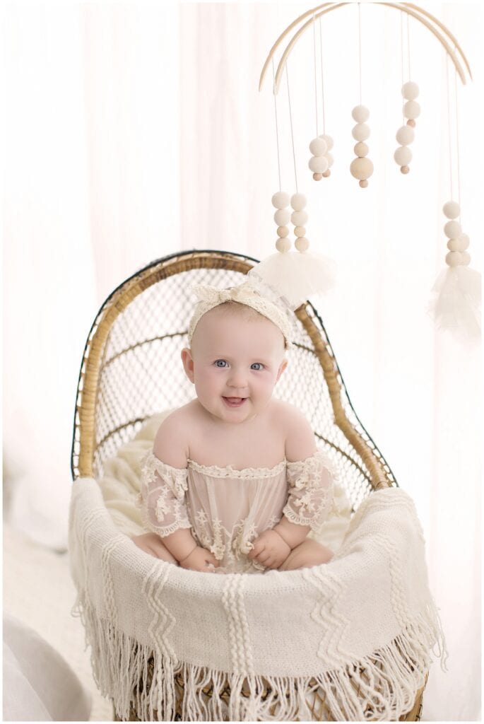 Smiling 6 month old baby girl sitting in a wicker bassinet with a cream lace outfit during her milestone photo session