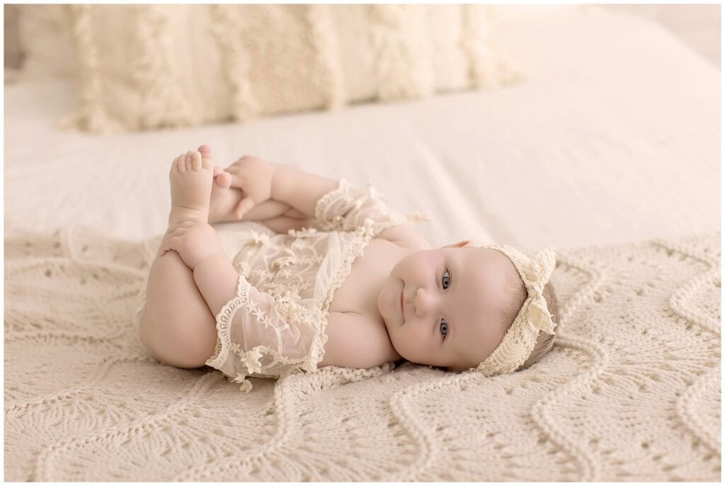 Six month old baby girl lying on a cream knit blanket holding her feet during her milestone photo session.
