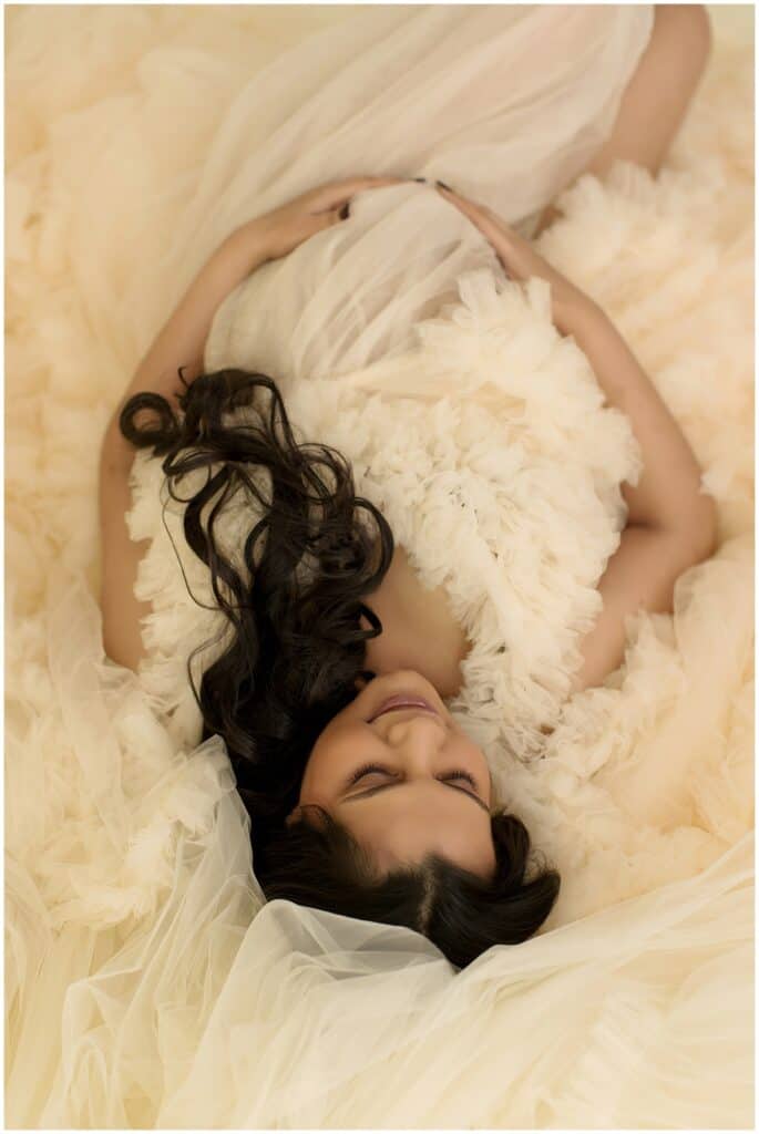 What to wear for your maternity photos- an expecting mom laying in a textured tulle backdrop with her baby bump