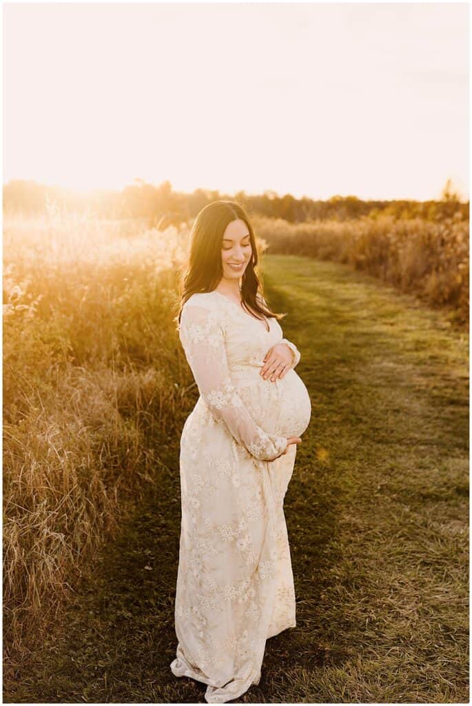 Golden hour outdoor maternity photo of expecting mother in cream lace dress walking through a Cleveland area field.