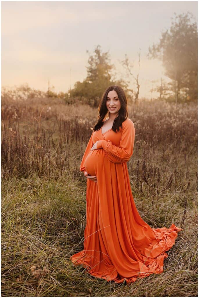 Spring outdoor maternity photo of pregnant woman in flowing orange dress standing in a field near Akron Ohio.