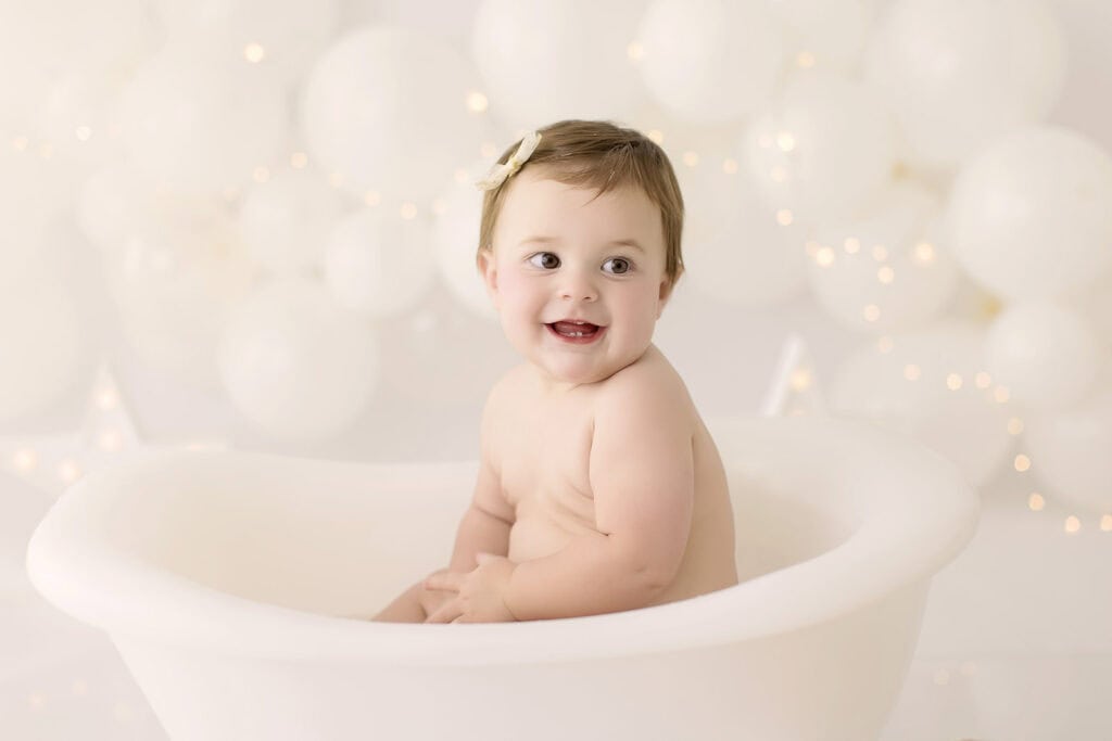 Baby girl smiling during a post cake smash bath session in a white tub with soft lighting